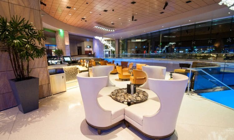 Modern lounge area with curved white seating, small round tables, tan chairs, and large windows overlooking a cityscape at night.