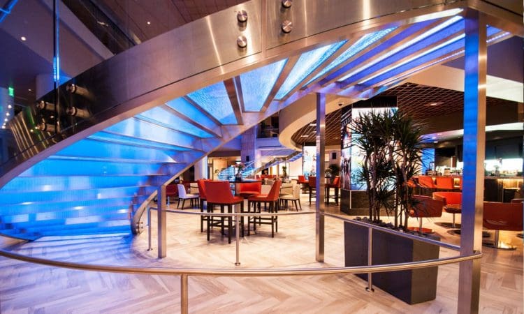 Modern restaurant interior with a curved, illuminated staircase, red chairs around tables, tiled flooring, and potted plants under a blue glow.