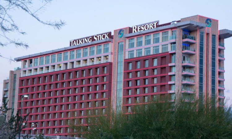 A multi-story hotel building with "Talking Stick Resort" signage on top, featuring red accents and surrounded by green foliage.