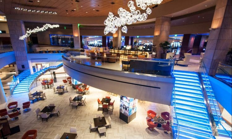 Spacious, modern hotel lobby with illuminated blue staircases, glass railings, round chandeliers, and tables with red and brown chairs arranged on a patterned floor.