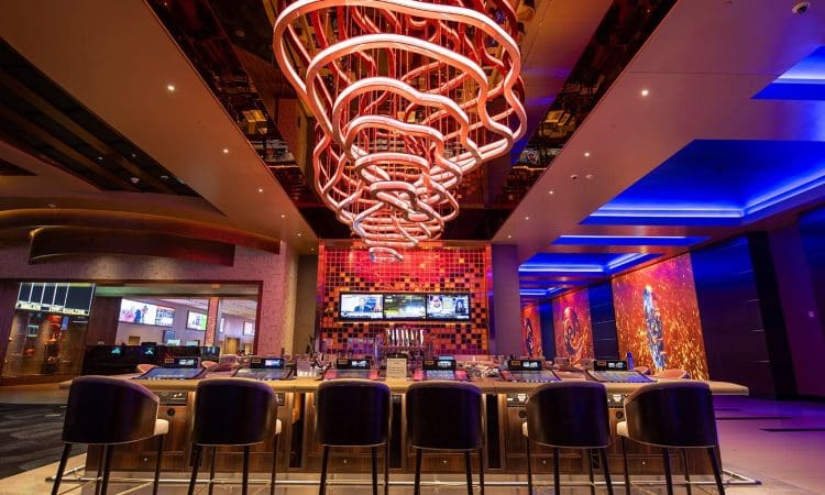 A modern casino bar with a large neon light fixture, bar stools, electronic gaming screens, and colorful lighting throughout the interior.