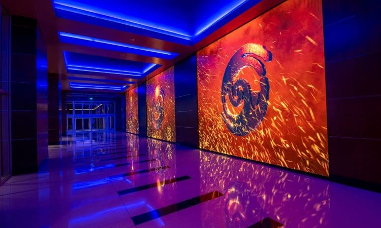 A hallway with blue overhead lighting and large digital screens on the wall displaying an abstract fiery design with swirling shapes.