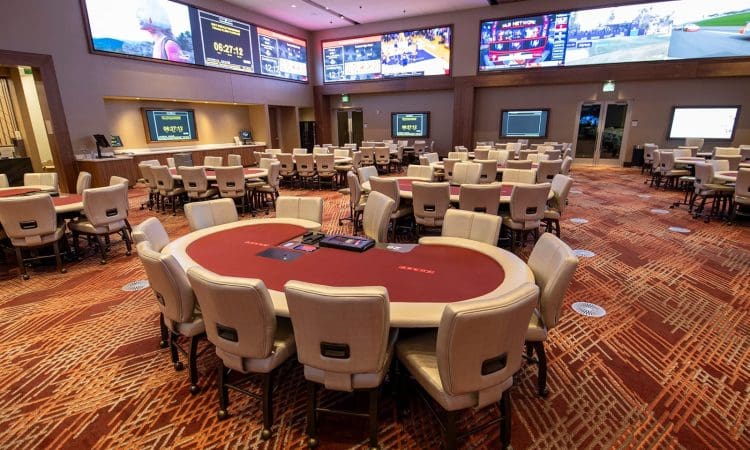 Spacious casino room with multiple round poker tables and chairs, large screens displaying sports events on the walls, and patterned carpet flooring.