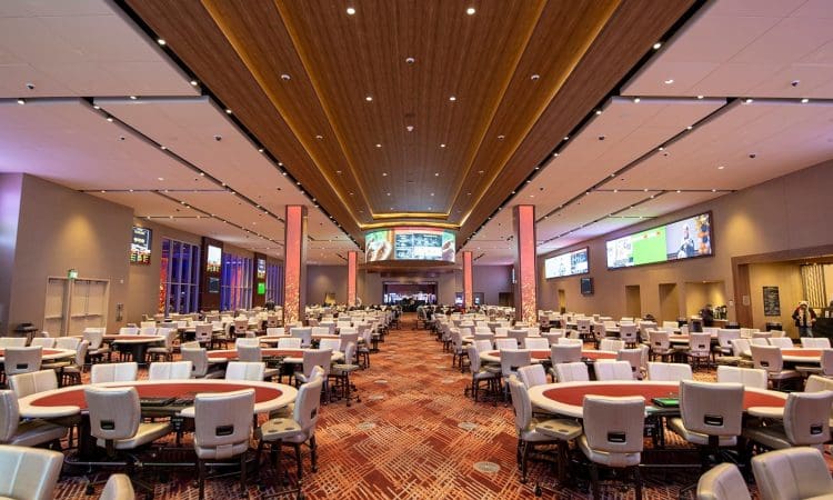 Wide view of a spacious casino poker room with numerous empty poker tables, modern lighting, and digital screens along the walls.