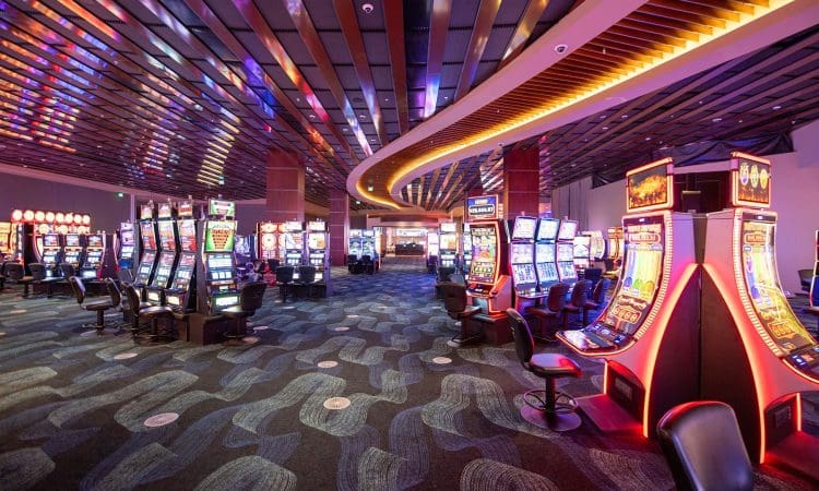 A spacious casino interior with rows of brightly lit slot machines, modern carpet, and a wavy ceiling feature; empty chairs are positioned at each machine.