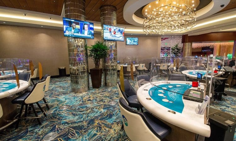 Casino gaming tables with plexiglass dividers, empty chairs, decorative lighting, TV screens displaying news, and patterned carpeted floor.