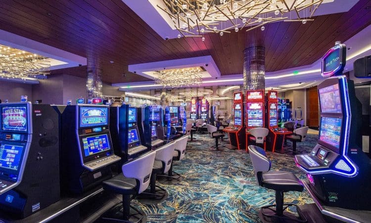 A brightly lit casino floor with rows of slot machines, empty chairs, patterned carpet, and decorative ceiling lights.