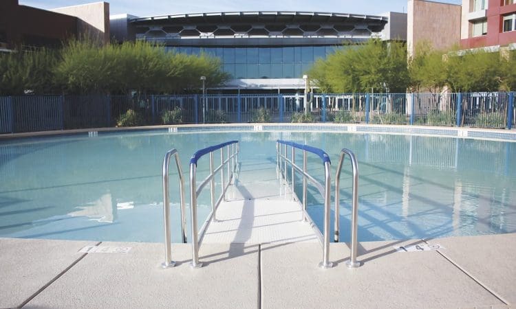A round outdoor pool with a central ramp and handrails, surrounded by a fence, with modern buildings and trees in the background.