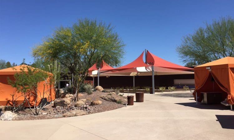 Outdoor area with orange tents, desert plants, and shade structures under a clear blue sky. Tables and chairs are set up beneath the largest canopy.