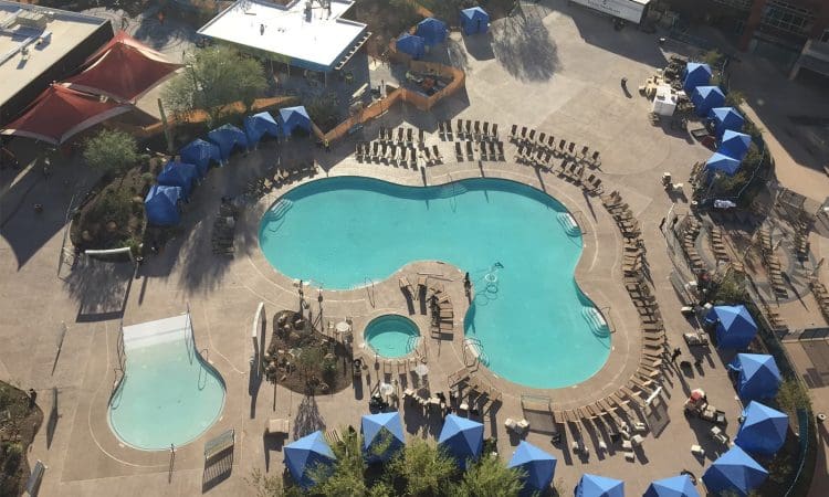 Aerial view of a hotel pool area with a large main pool, a smaller round pool, a shallow wading pool, and blue cabanas arranged around the deck.