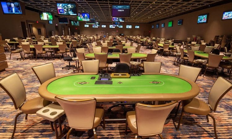 A large, empty casino poker room with rows of green poker tables, tan chairs, overhead lights, and multiple televisions on the walls.