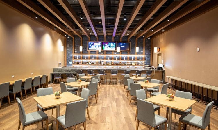 Spacious modern restaurant with wooden tables and gray chairs, facing a long bar with multiple TVs and bottles displayed on shelves behind it.