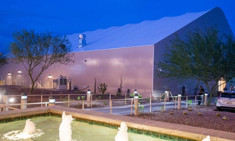 A modern building with a white exterior is seen at dusk, with a fountain in the foreground, desert landscaping, and a few people walking near the entrance.