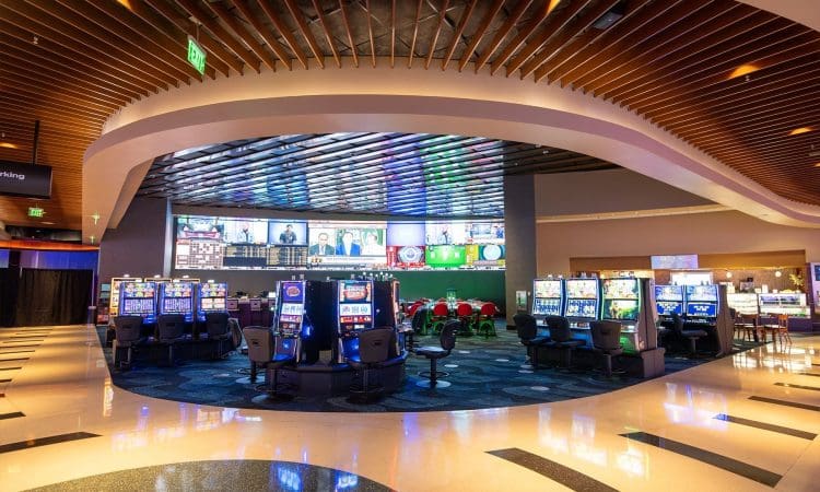 Casino interior with rows of slot machines, digital screens displaying sports, and gaming tables under a curved, modern ceiling.
