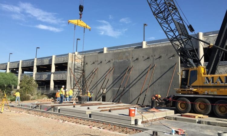 Construction workers use cranes and equipment to install concrete panels at a building site near a multi-level parking structure under a clear blue sky.