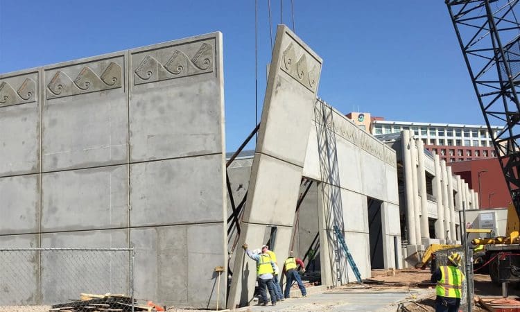 Construction workers guide large concrete wall panels into place with a crane at a building site under clear blue sky.