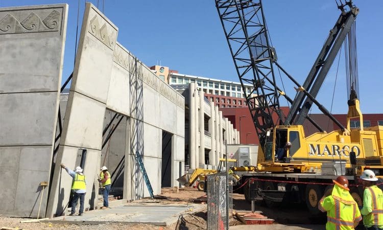 Construction workers guide a large concrete wall panel into place with the help of a Marco crane at a building site under clear blue sky.