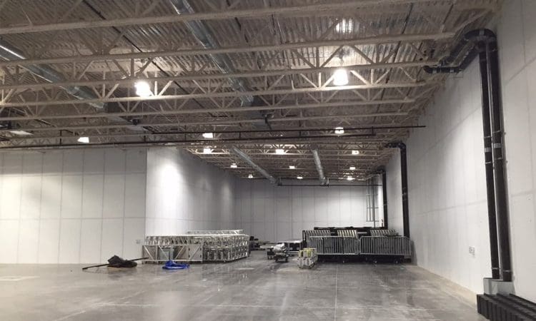 Large empty warehouse space with high ceilings, exposed beams, and stacked metal structures and equipment along the back and right walls.