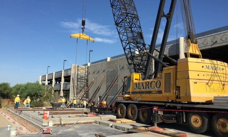 Construction workers use a large yellow crane to lift and position concrete panels near a partially built structure under a clear blue sky.