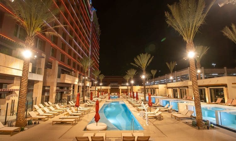 Outdoor hotel pool area at night with lounge chairs, red umbrellas, tall palm trees, and a multi-story building illuminated with colorful lights in the background.