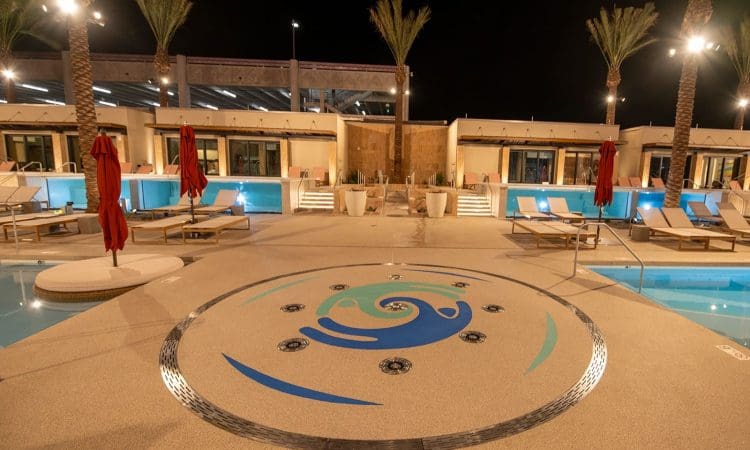 Outdoor swimming pool area at night with lounge chairs, red umbrellas, palm trees, and a circular decorative design on the pool deck. Modern building and pool lights in the background.
