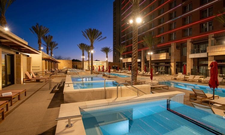 Outdoor pool area at a modern hotel, illuminated at dusk, with palm trees, sun loungers, and red umbrellas surrounding several pools and hot tubs.