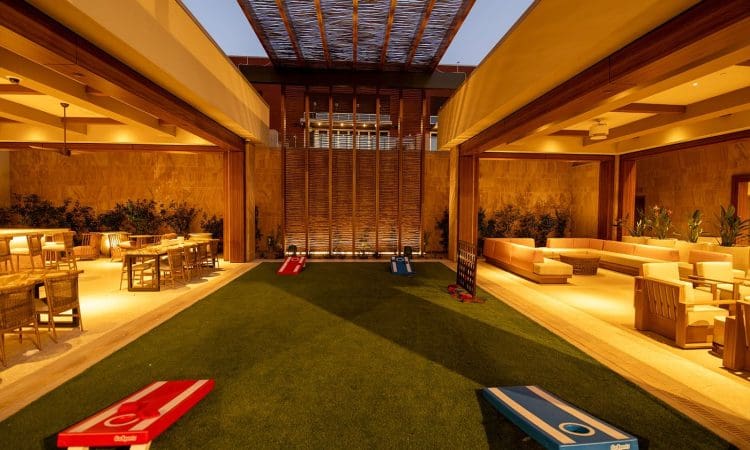 A modern open-air lounge with seating areas on both sides and two cornhole games set up on a central artificial grass area, lit by warm ambient lighting.