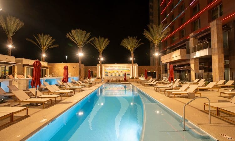 A modern outdoor pool at night, surrounded by lounge chairs and red umbrellas, with palm trees and a lit building in the background.