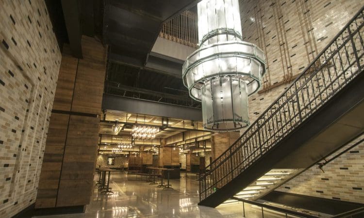 Modern interior with exposed brick walls, metal staircase, polished floors, and a large cylindrical glass chandelier hanging from the ceiling. Tables and chairs are arranged in the background.