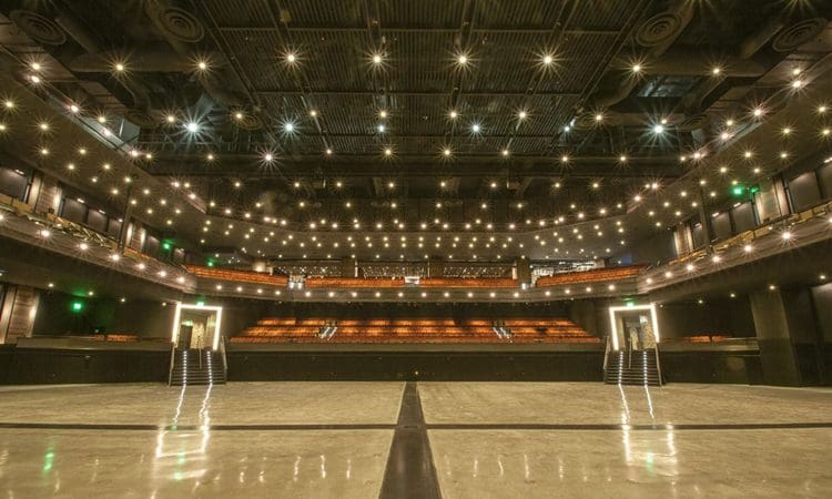 A wide view of a modern, empty theater auditorium with rows of orange seats, two balconies, and ceiling lights illuminating the space.
