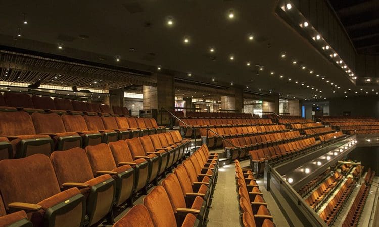 Rows of empty, brown theater seats in a modern auditorium with overhead lights and a view of the stage area to the right.