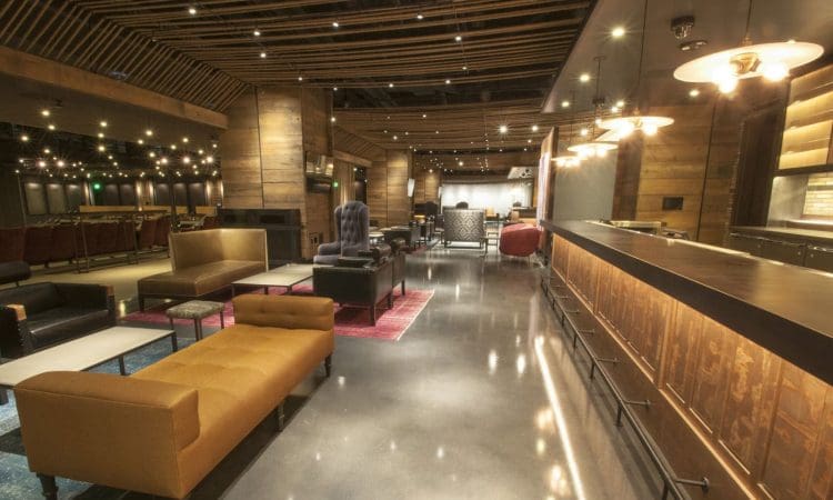 Spacious modern lounge with wood accents, upholstered seating, a long bar, polished concrete floor, and warm overhead lighting.