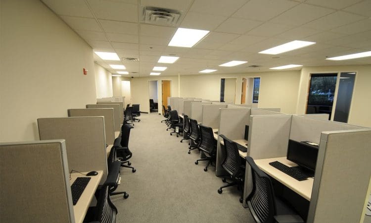 A modern office with rows of empty cubicles, each equipped with a computer workstation and black office chairs, under bright ceiling lights.