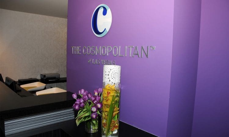 Reception desk at The Cosmopolitan of Las Vegas with a purple wall, company logo, fresh flowers, and a decorative vase.