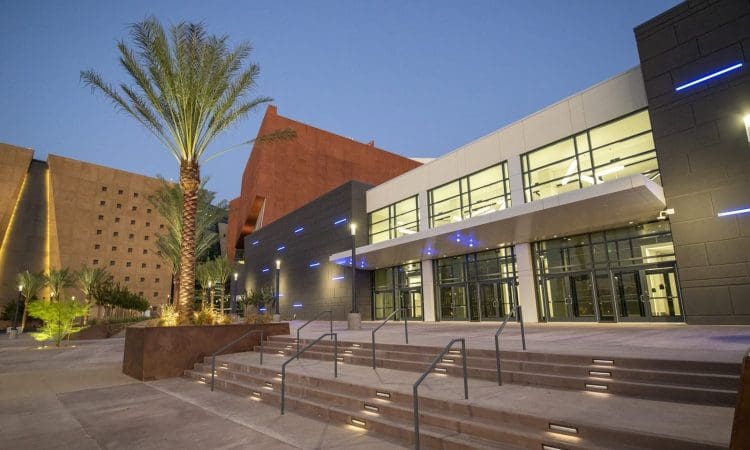 Modern building with large windows, illuminated steps, and a palm tree in front, photographed at dusk with clear skies.