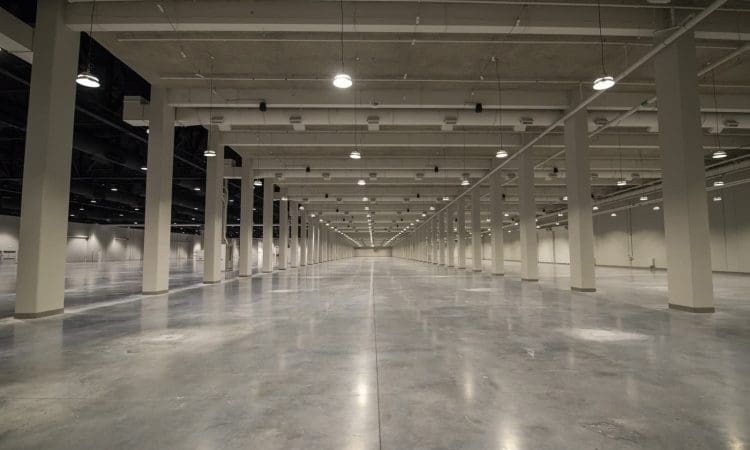 Large, empty warehouse with polished concrete floor, numerous ceiling lights, and rows of support columns extending into the distance.