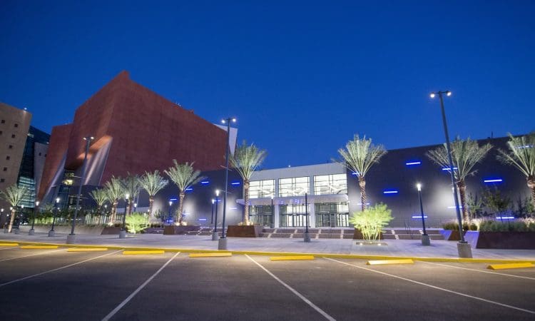 Modern building with illuminated blue accents, palm trees, and a mostly empty parking lot in the foreground at dusk.