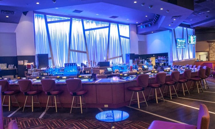 A modern bar with red chairs surrounds a counter, illuminated by blue lighting and draped curtains, with bottles and screens visible behind the bar.