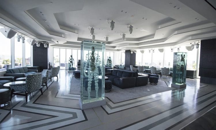 Spacious modern lounge with large windows, patterned chairs, sofas, geometric flooring, and two tall glass sculptures in the center of the room.