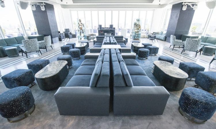 Spacious, modern lounge with blue sofas, patterned armchairs, round ottomans, and marble coffee tables, set in a room with large windows and city views.