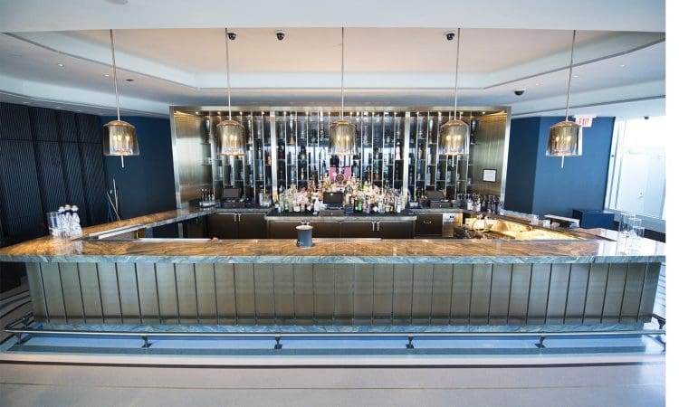 Modern bar with a marble countertop, backlit shelves displaying bottles, and pendant lights hanging from the ceiling in a sleek, minimalist setting.
