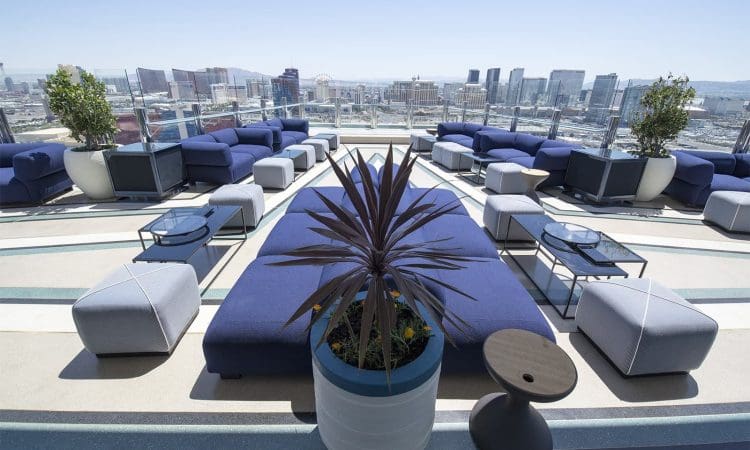 Rooftop lounge area with blue cushioned seating, tables, potted plants, and a city skyline visible in the background under a clear sky.