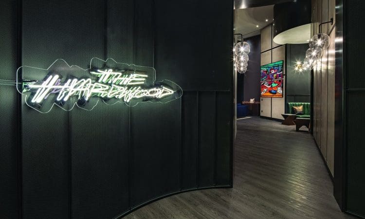 Dimly lit modern hallway with neon sign reading "The Harlewood," dark walls, wooden floor, hanging lights, and colorful framed artwork in the background.