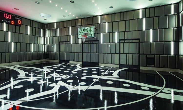 Modern indoor basketball court with a graphic black-and-white floor design, padded walls, digital scoreboard, and ceiling lights. A basketball hoop is mounted on one wall.