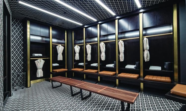 Modern locker room with dark wood lockers, beige robes hanging, neatly folded towels, a wooden bench, and patterned black-and-white flooring and walls. Bright ceiling lights illuminate the space.