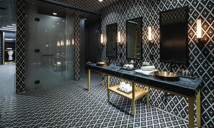 Modern bathroom with black and white geometric patterned walls and floor, double sinks with gold accents, mirrors, wall lights, and folded towels on shelves underneath.