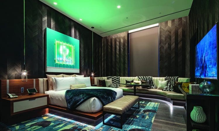 Modern hotel room with a large bed, sectional sofa, wall-mounted TV, abstract art, and green accent lighting creating a contemporary atmosphere.