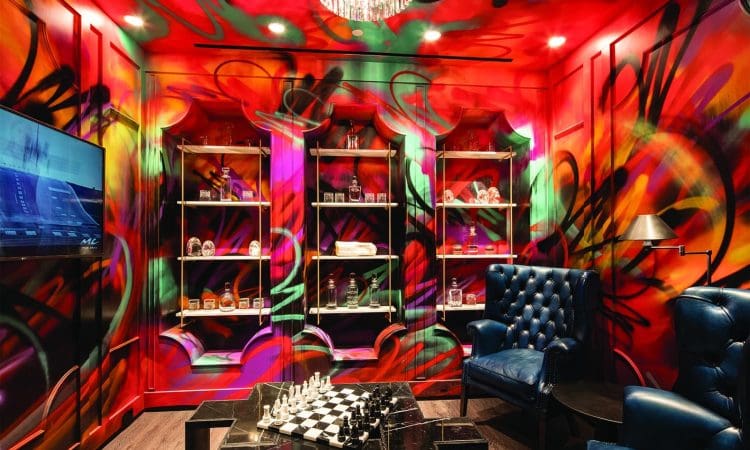 A lounge with vividly painted graffiti walls, glass shelves with bottles, blue leather chairs, a chessboard table, and a chandelier overhead.