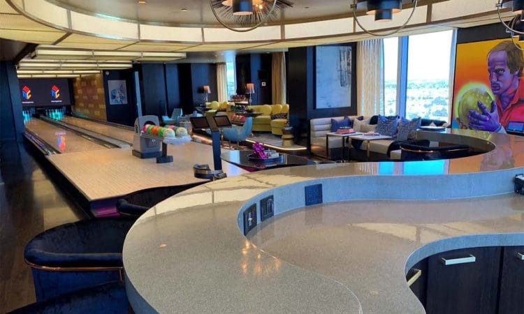 A modern lounge with a bowling alley, bar counter, couches, colorful bowling balls, and a large painting of a person holding a bowling ball on the wall.