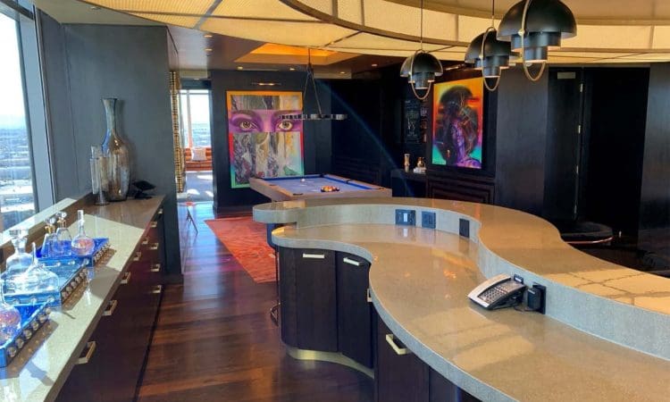 Modern lounge with curved stone countertop, pool table, bold colorful art on dark walls, pendant lights, and city view through large windows.